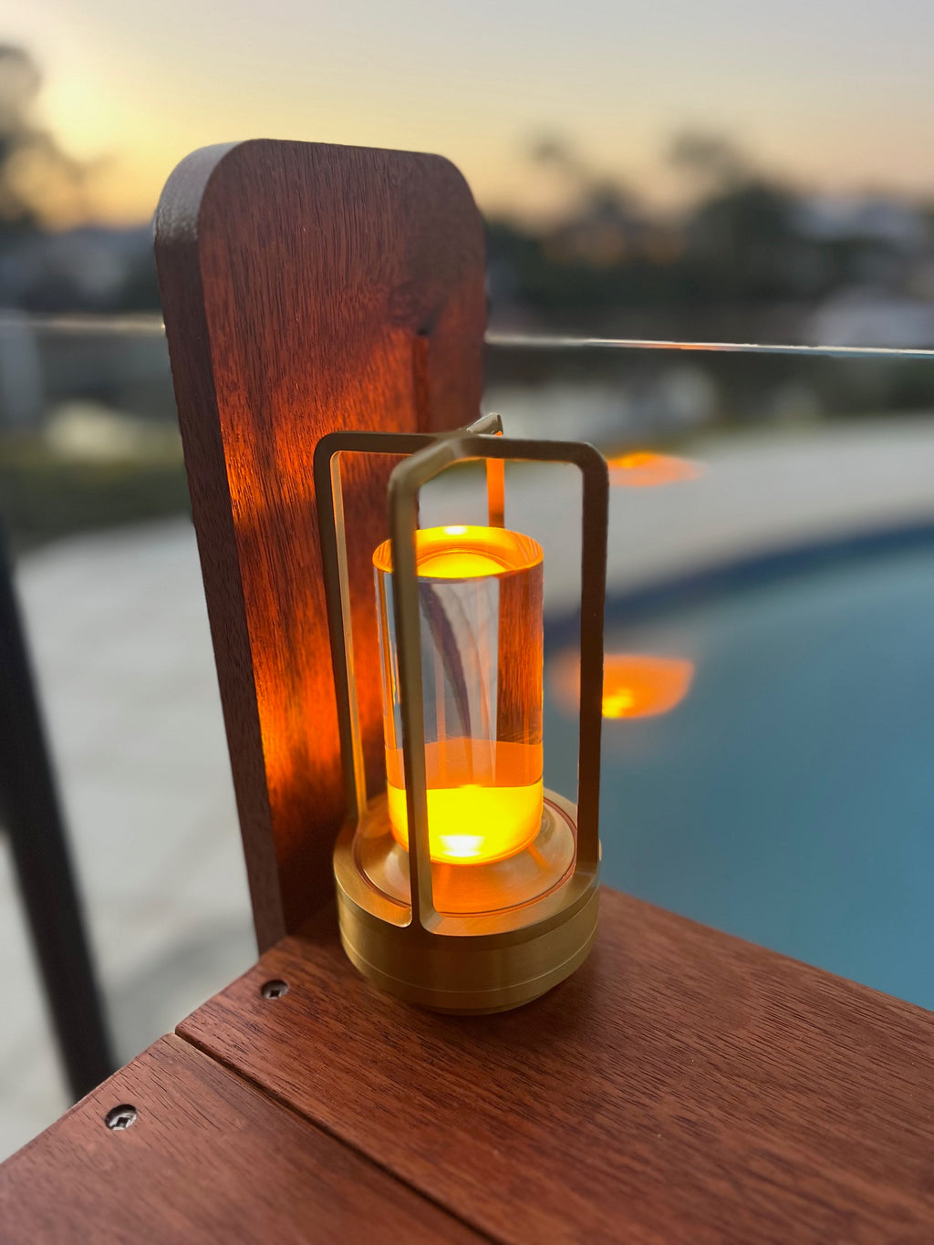 Wooden lantern with glowing interior on a railing by a poolside at sunset.