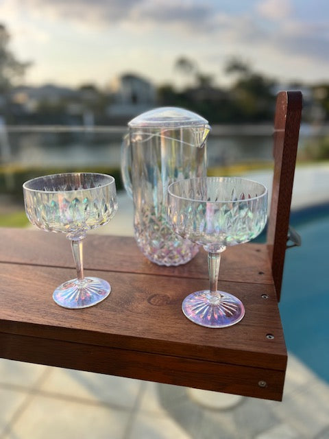 Two iridescent cocktail glasses and a pitcher on a wooden ledge with a blurred outdoor background.