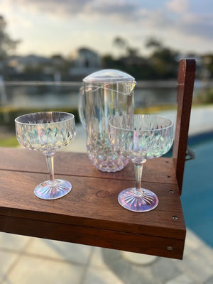 Two iridescent cocktail glasses and a pitcher on a wooden ledge with a blurred outdoor background.